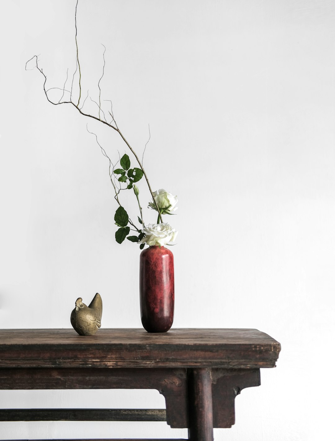 Minimalist Chinese style, a simple and elegant space composition. A dark red ceramic vase sits on an old wooden table containing one white rose. Green branches and a golden bird figurine are beside the flower stand against a white background. The high definition photography features soft lighting, natural light, and warm tones with clear details of the natural elements, delicate textures, and a natural atmosphere. –ar 97:128