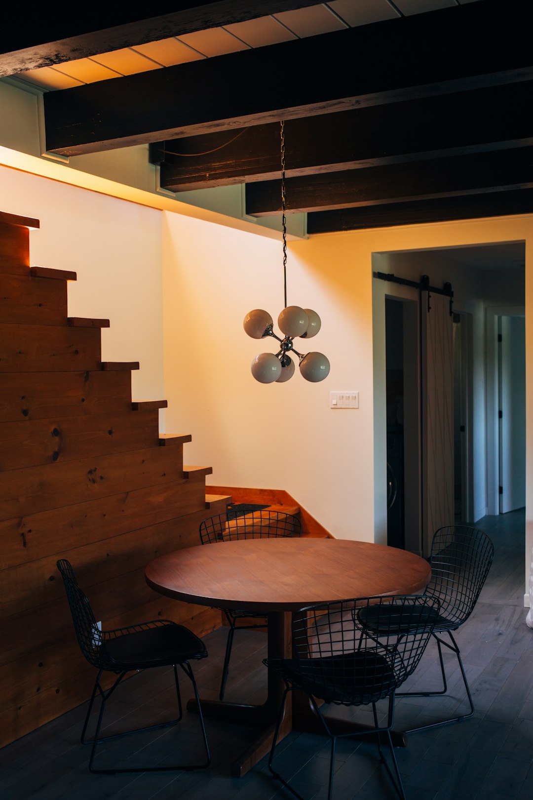 Small dining room in a tiny house, with a round table and black chairs, warm lighting, a wooden staircase on the side of the wall, a simple ceiling with dark wood beams, light grey walls, a modern pendant chandelier, a photo in the style of Canon EOS R5 F2 ISO400 with ultra detail. –ar 85:128