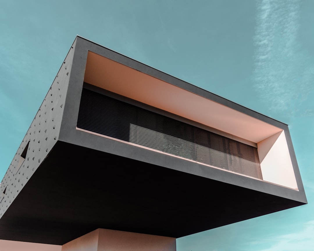 A modern minimalist house with a flat roof and an exterior balcony on the right side of its two floors, featuring dark gray concrete and light white metal materials. The photo was taken from directly below looking up at it, with a clear blue sky in the background. A high angle perspective, closeup shot focusing on details such as textures, shadows, and highlights to highlight the architectural design. A Canon EOS R5 camera was used for its sharpness and ability to capture detail.