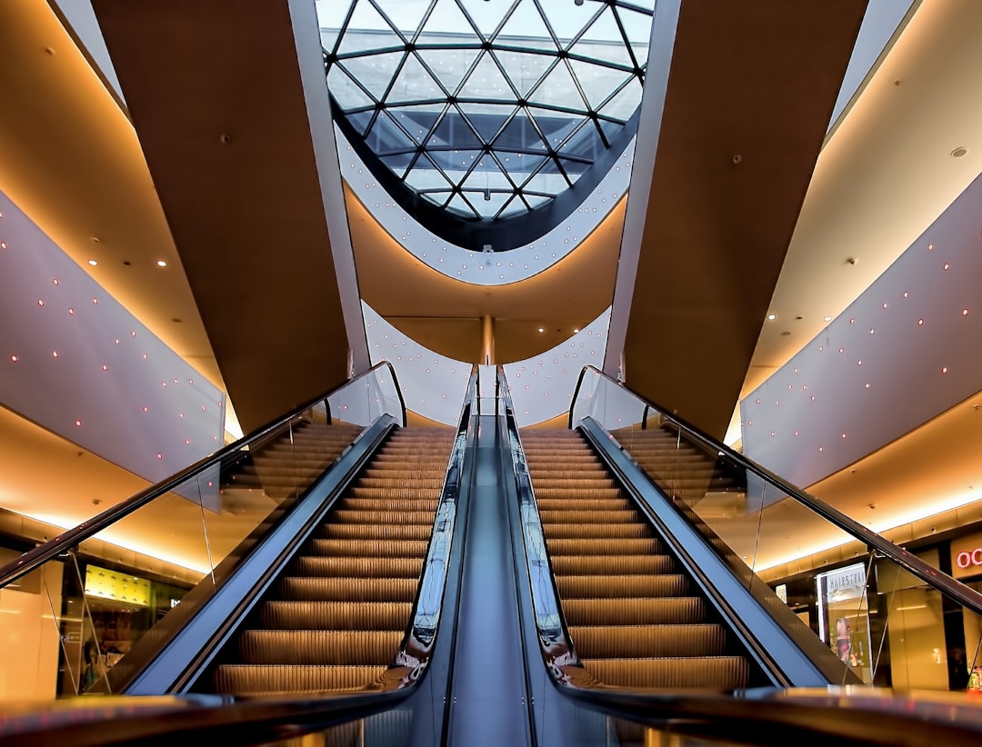 A photo of an indoor shopping mall with two escalators, a glass dome ceiling above the stairs and light brown carpet on both sides of each staircase. The overall atmosphere is modern, with soft lighting illuminating the interior space. In front of it lies another staircase leading to ground level, adding depth to its architecture. This scene captures a wideangle view from directly below looking up at the entire structure, showcasing a spacious environment with a contemporary aesthetic in the style of a modern architect. –ar 128:97