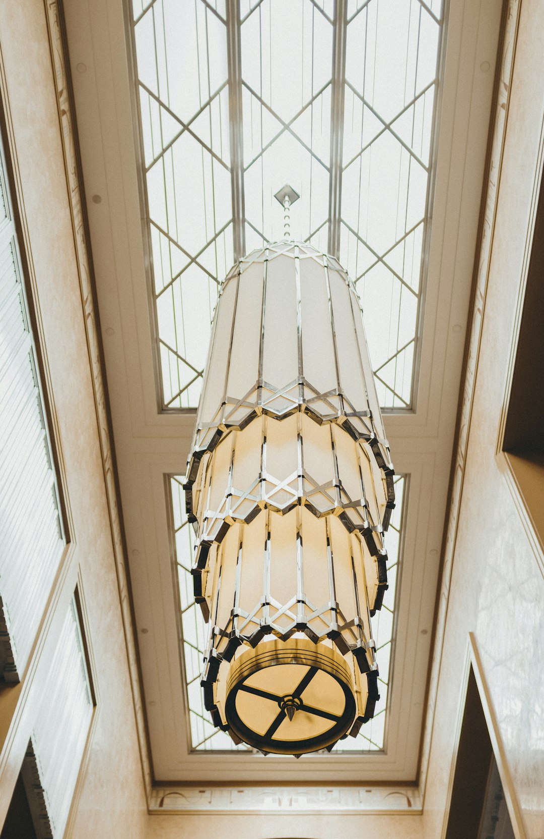 A large art deco chandelier hanging from the ceiling of an entrance hall, designed in the style of Dunand. The glass and fabric shade is white with black trim. Use natural light to highlight its elegant design. A high angle perspective capturing the full scale of the pendant in front of tall windows. Soft lighting creates a warm glow around it. A closeup shot focusing on intricate details such as geometric patterns or textures within the lamp’s frame. –ar 83:128