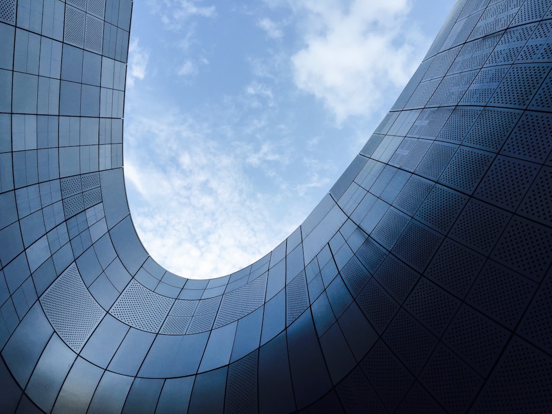 A photo of the exterior wall, curved building made from steel with blue glass windows, low angle shot looking up at sky, modern architecture, minimalistic, steel facade with architectural details, curved panels, blue sky and clouds in background, daylight, architectural photography, archdaily, architecture
