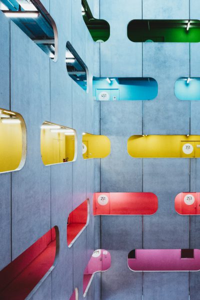 A colorful, playful display of futuristic letter boxes with numbers on them hanging from the ceiling in an office space. The background is a grey and blue concrete wall.