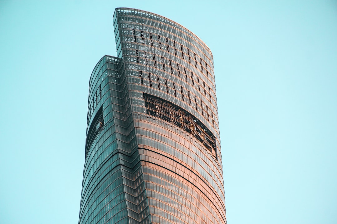 A tall, slender skyscraper with glass facades against the blue sky. The building has curved lines and is decorated in shades of pink and orange. It stands alone on an empty urban landscape. There is no one around or any other buildings nearby. Captured from below using a Canon EF lens at an f/2.8 aperture setting in the style of no particular artist.