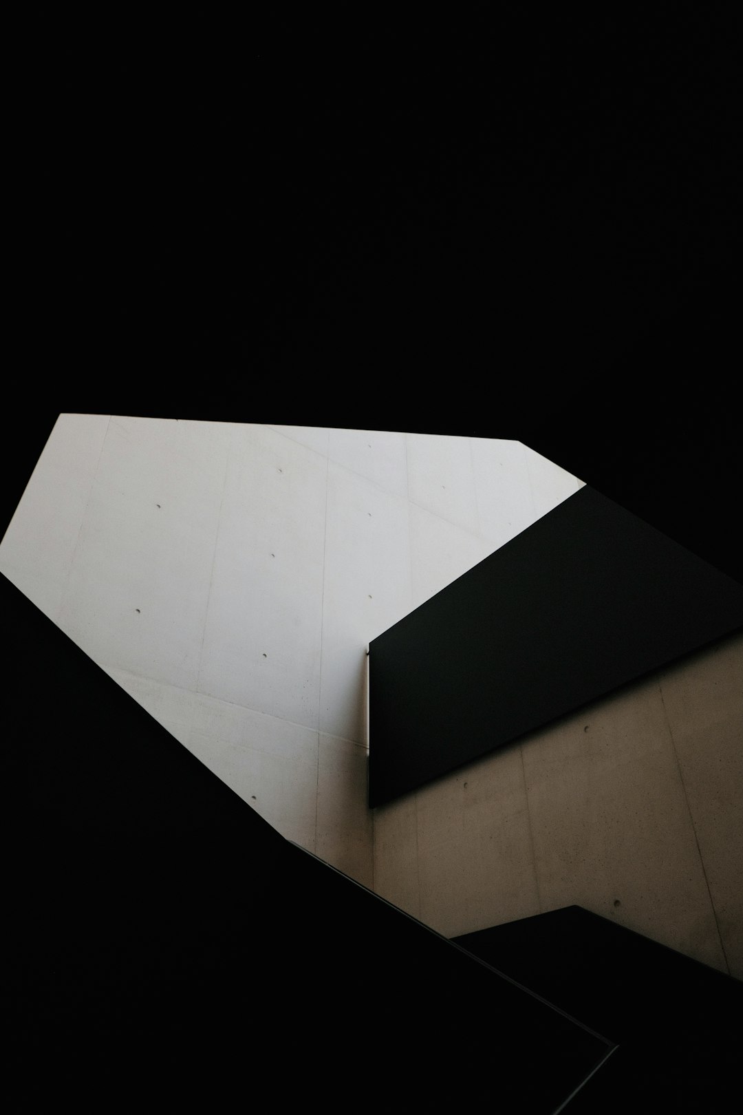 A photo shows the inside corner of an angular white and black building, taken from above. Light is coming in through one side of the space creating shadows on the wall behind. It is a minimalistic scene with only two colors. The photo was shot using Kodak Portra film stock, giving it a cinematic quality in the style of the film.