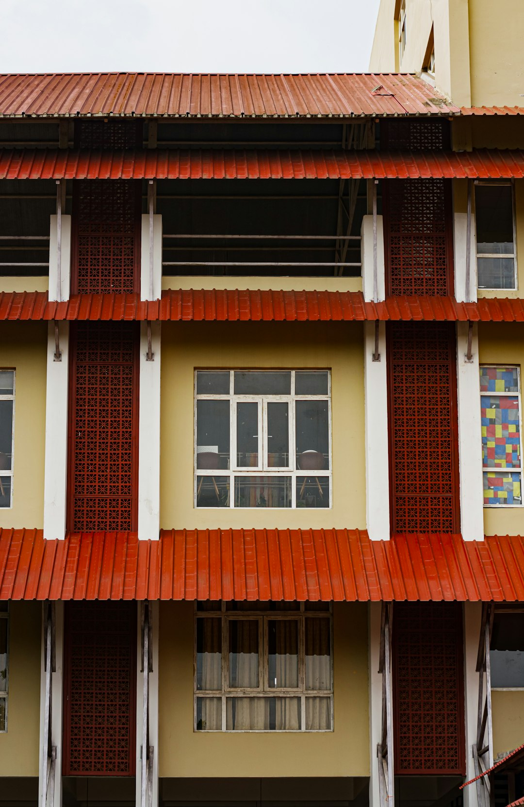 a school building in singapore with red and white tiles on the roof, photo taken from street level, large windows, modern architecture, front view, white walls, wooden columns, windowsill decoration, multilevel structure, a young girl is standing at one of its window, day time, light yellow sky, red bricks and wood details on wall –ar 83:128