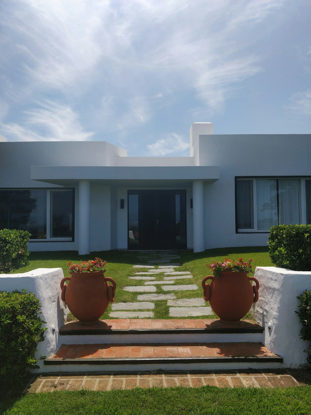 A simple, white modern house with terracotta pots and a front door in the center of Cartagena’s coast. The entrance has two steps leading to an open space on one side of which is located a red clay pot filled with flowers. On both sides there are green hedges. In the background you can see a clear blue sky and calm sea. The photo was taken in the style of an iPhone 7. –ar 3:4