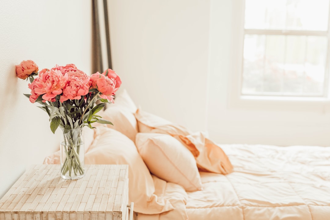 a minimalistic bedroom with peach colored flowers in vase on bedside table, white walls, clean and tidy room, window light coming from the right side of picture,