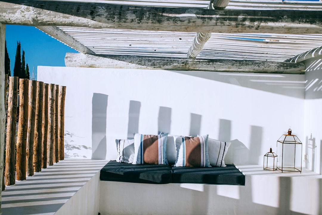 White walls, terracotta wood and concrete accents, black cushions on the roof terrace of Mykonos island in Greece with blue sky, sun rays, shadowplay, rustic architecture, interior design photography, bohemian style decor. –ar 128:85