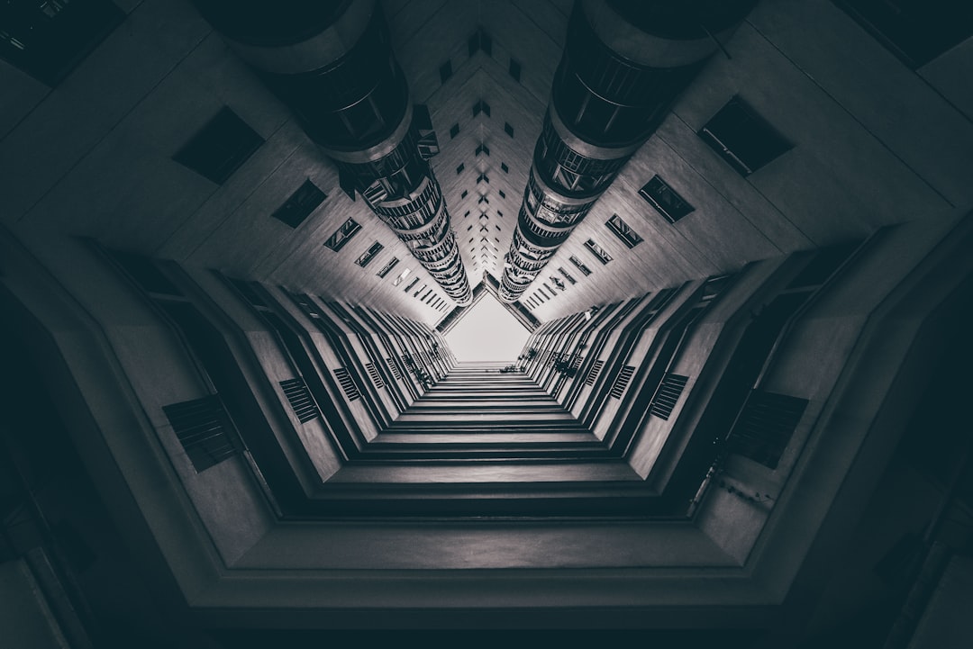 Abstract architecture photography of a high rise apartment complex. The symmetrical composition looks up from the ground through a narrow hallway towards the ceiling with large columns on either sides. It has a dark and moody style with high contrast and a monochrome color scheme. The cinematic lighting adds to the mood.