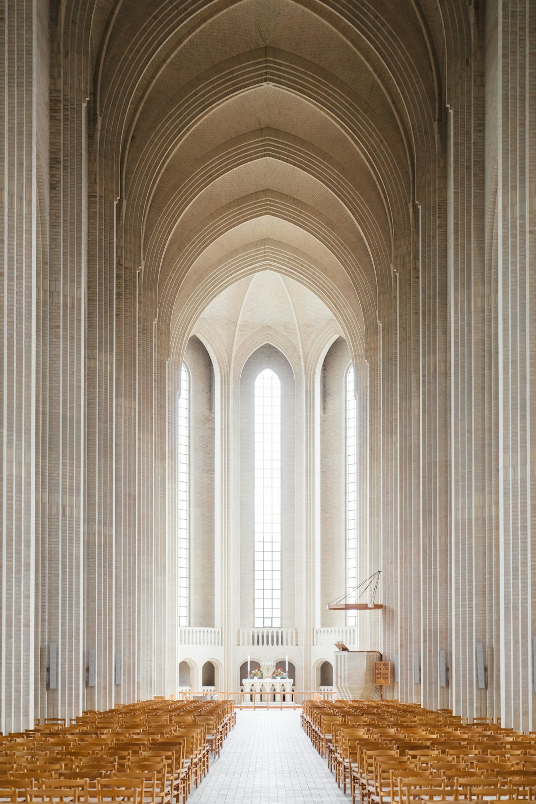 The interior of an impressive cathedral in Denmark designed in the style of Irving Pei and [Tadao Ando](https://goo.gl/search?artist%20Tadao%20Ando) in a modernist style. The space is large with high arched ceilings. There are no stuccos on the walls or columns, only very thin white concrete slabs. In front there are rows upon rows of wooden pews leading to the altar at the far end of the room. A single long window brings light into the room from outside. –ar 85:128