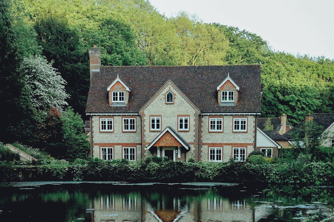 A picturesque English country house nestled amidst lush greenery, with a serene river flowing in front of it and reflecting the charming architecture. The photo captures the style of Canon EOS R5 T2i, with an unsplash photography style of minimalism, cinematic natural light, highly detailed, high resolution.