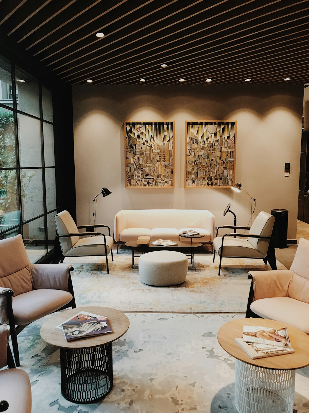 A modern hotel lounge area with three paintings hanging on the wall, sofa and chairs, coffee tables, magazine sofas, and natural light coming in from large windows. The walls have dark beige tones and wooden beams above them, creating an atmosphere of luxury and comfort. There is also some artwork on display that adds to its artistic ambiance. This space exudes tranquility and elegance for guests’ relaxation time during their stay at our vacation DirectSheetXl4LkMj .836x705_2 logo