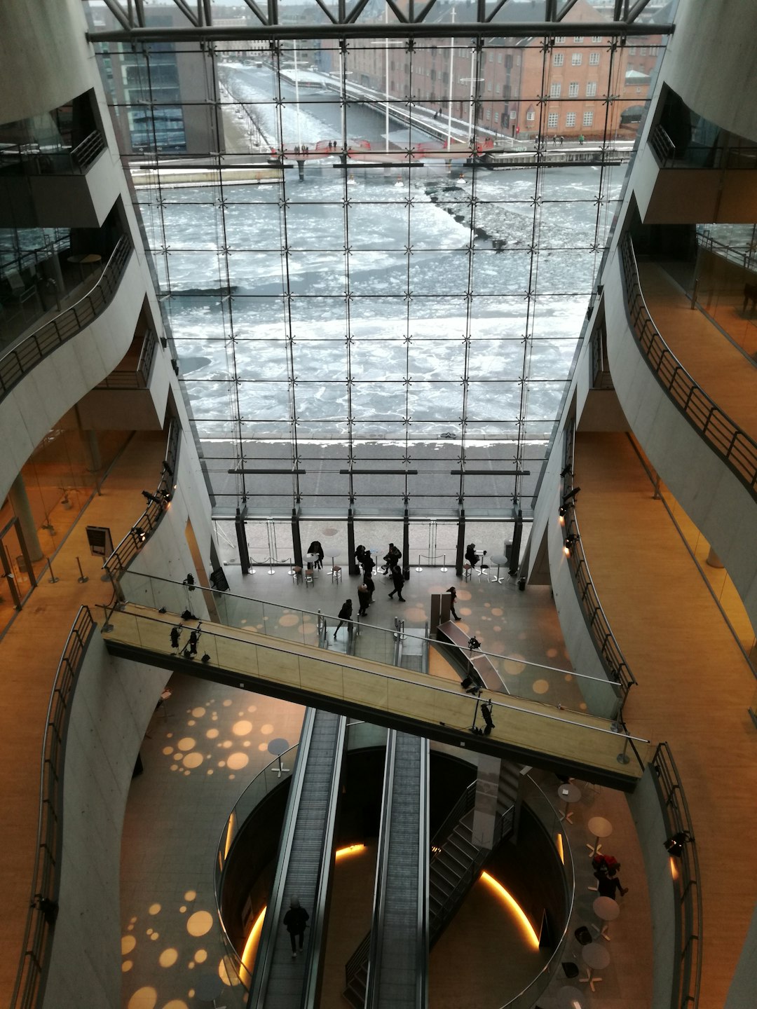 a photo of the interior space in new postmodernism style, with large glass windows overlooking an icy river and people walking on it. The building has three floors and is surrounded by modern architecture. There’s an indoor staircase leading to different levels inside the structure. On one side there’s another tall window that reveals part of city buildings outside. People can be seen moving around within the scene. It feels bright and airy with natural light streaming through the ceiling.