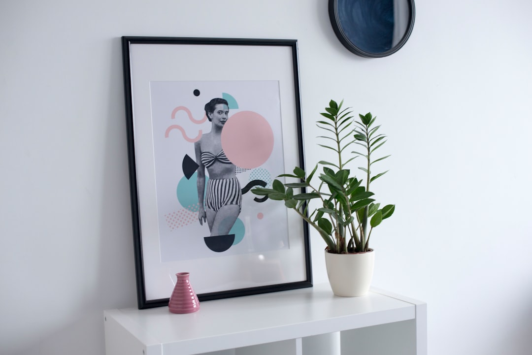 A framed collage art print on white wall with black frame, modern abstract shapes of female figure in fashion outfit, standing pose, potted plant and pink vase on side table, in style by light blue and soft pastel green color palette, in the background is clean plain white wall, aesthetic picture frame mockup, high resolution –ar 128:85