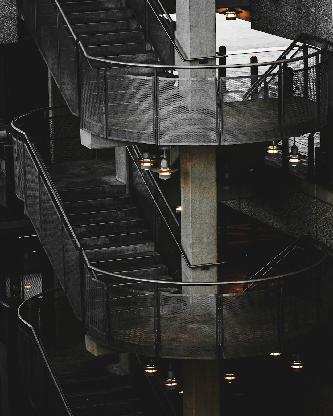 Dark gray concrete spiral staircase, concrete walls with glass windows on the sides of each step, dark metal hand railings, light bulbs hanging from ceiling, photo taken by Sony Alpha A7 III –ar 51:64