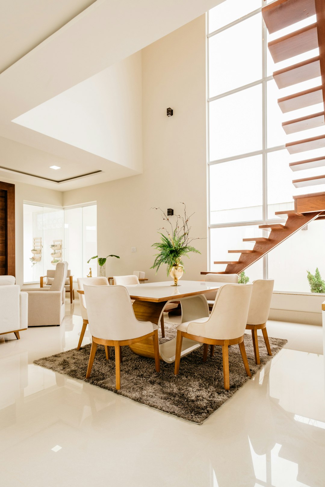 modern dining area in the middle of a large living room, with white walls and ceiling, and a wooden staircase on the side. The floor is white marble with some carpeted areas. There is a glass window allowing natural light from big windows with a neutral tones color palette. The shot is from a high angle, showing an interior home design in the contemporary style. –ar 85:128