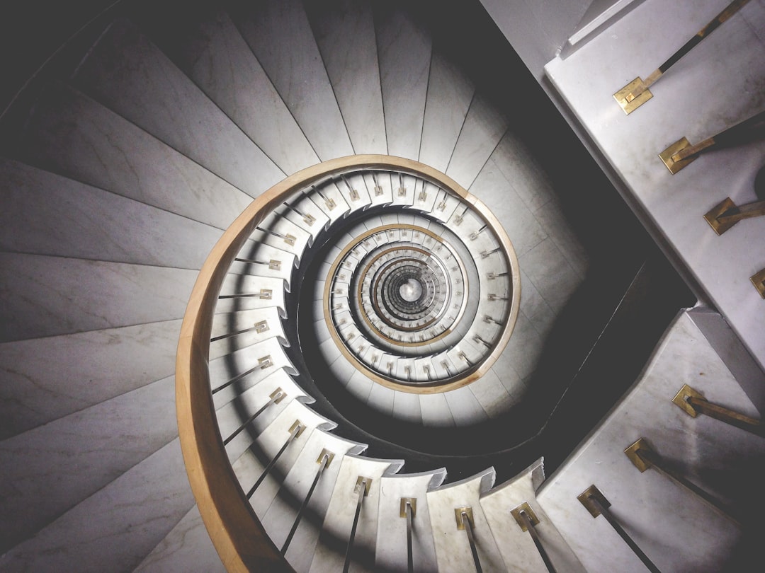 A spiral staircase with white marble steps and brass accents is viewed from above. The camera captures the circular pattern of the stairs in muted tones against a light background. A sense of elegance is captured as well as the impression one would feel if stepping down these stairs. This photo was taken in the style of Canon EOS R5 using a macro lens.