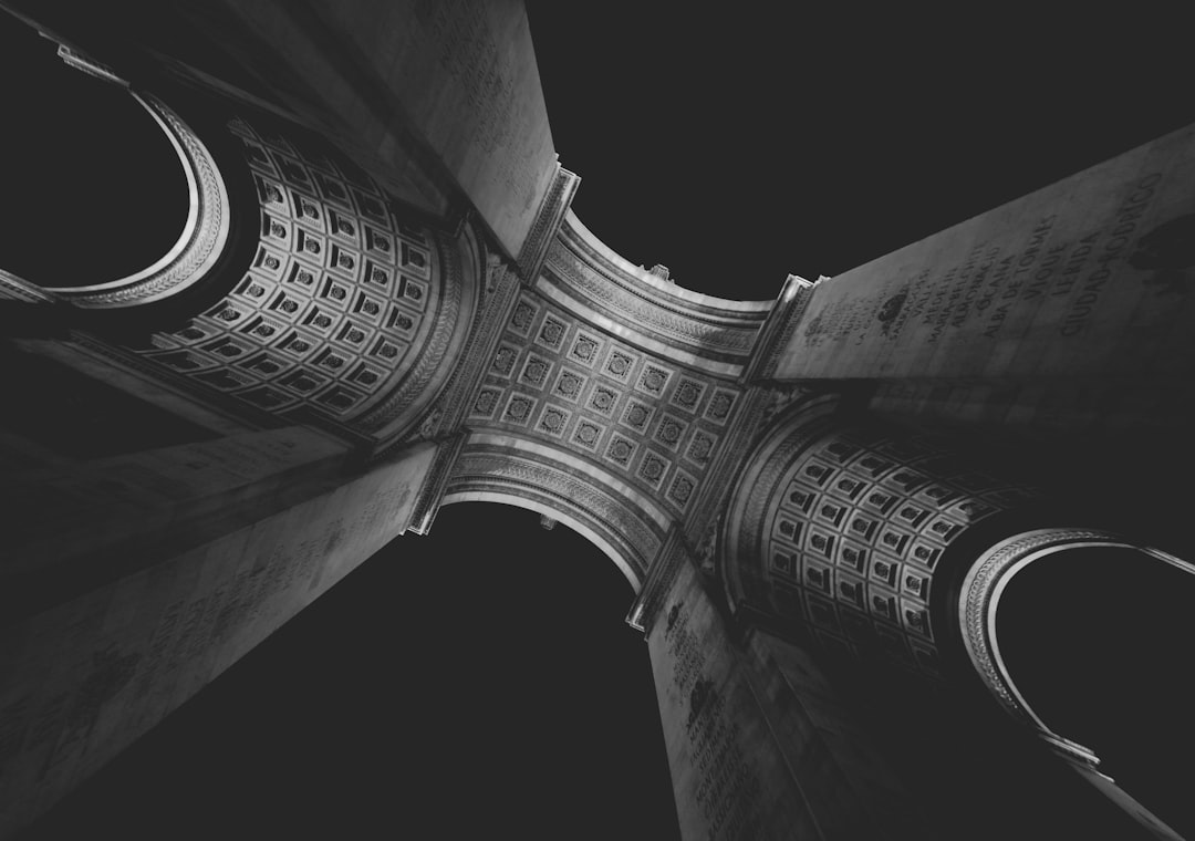 A black and white photo of the arches at Place de la Triomphe in Paris, shot from below looking up, symmetrical, low angle, cinematic, high contrast, dark, moody, with a film grain effect. The photo is in the style of a dramatic, low-light composition that enhances the architectural lines and shapes. –ar 64:45