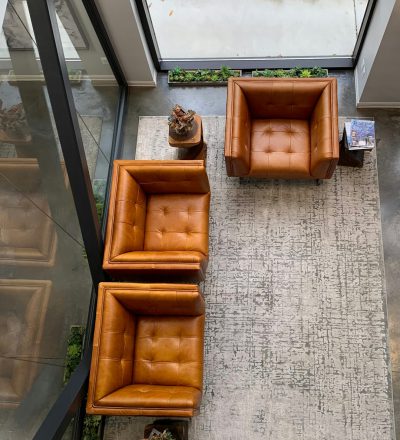 Photograph of three leather armchairs arranged in an L shape, viewed from above, placed on the carpeted floor of a modern living room with glass walls and concrete floors. The chairs have brown tones and there is also some greenery outside. On one side of each chair sits a small vase containing flowers or plants. There's another object next to them that could be a photo frame. In front of these chairs lies a gray rug with white designs in the style of an abstract artist.