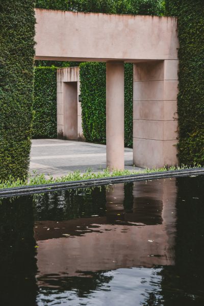 A minimalistic photo of an architectural garden, with symmetrical hedges and a water feature leading to the entrance gate of pink stucco walls, reflecting in still waters. The scene is captured from eye level, emphasizing symmetry and the contrast between natural greenery and modern architecture. Inspired in the style of [Peter Zumthor](https://goo.gl/search?artist%20Peter%20Zumthor)'s outdoor design for Aman shot on Fujifilm Provia film.