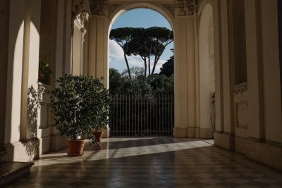 A view of the garden from inside an old palace in Rome, the entrance is through an arched door with large windows on each side. It's sunny and there's a tree outside behind a black metal fence. The floor has a patterned tile design. There are some plants next to the doors. The lighting should be soft and natural, in the style of an old palace.