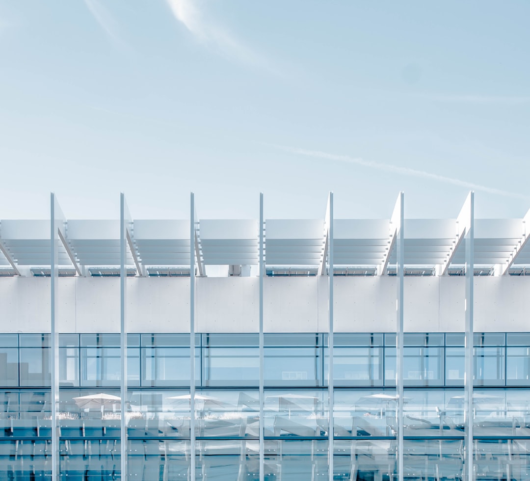 A white minimalist architecture with large columns and roof beams, featuring glass windows on the side of an airport building under a clear blue sky. The scene includes modern furniture in front of it, creating a sense of calmness and simplicity, with sunlight casting long shadows over the structure. This is captured from above using a Sony Alpha A7 III camera with a Zeiss Batis lens at a f/2.8 aperture setting, in the style of minimalist architecture. –ar 32:29