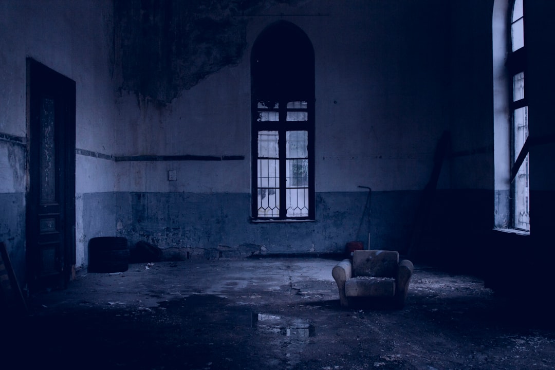 a dark abandoned room with a window, the floor is made of concrete and there’s an armchair in one corner, the walls have blue tones, the atmosphere is gloomy, the windows light up a small part of it, photo taken by Canon camera, –ar 128:85