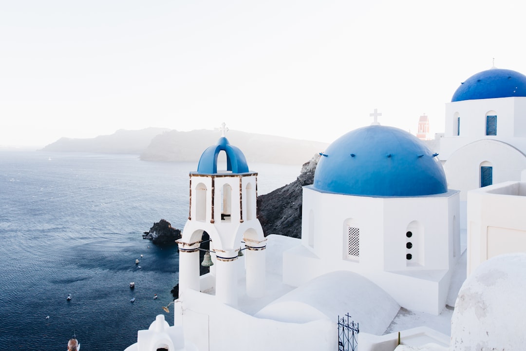 A photo of white and blue buildings in Santorini, Greece with the sea behind them. The sky is clear, and there is some mist on top of one church dome. It feels peaceful and serene. There are no people or cars visible, only the beauty of nature around us. This shot would be perfect for travel and vacation marketing materials in the style of peaceful and serene landscapes. –ar 128:85