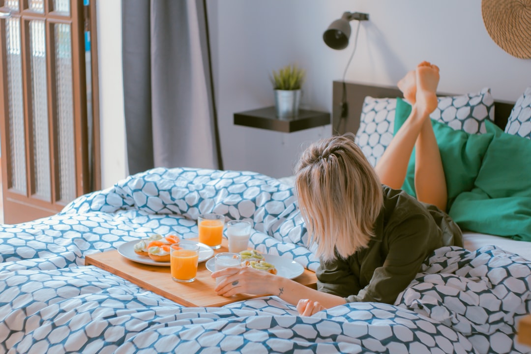 A woman lies on her bed with blue and white patterned sheets, enjoying breakfast in the morning light of natural sunlight. The wooden tray is filled with orange juice, fresh fruit salad, slices of bread for toast, and pastries. She has blonde hair tied back into an elegant low bun, wearing comfortable green pajamas. Her feet rest atop the bedside table near stylish black lampshades, creating a cozy atmosphere that reflects relaxation and tranquility during the early afternoon.