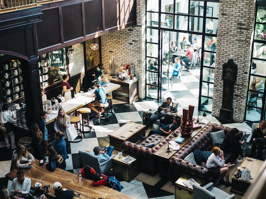 An overhead view of an open-plan office space with brick walls and large windows, with bustling activity going on around the room as people interact in lounges, coffee shops, and on couches, with a bar vibe and cool, hipster vibes. The high resolution photography captures the scene in the style of modern, urban photography.