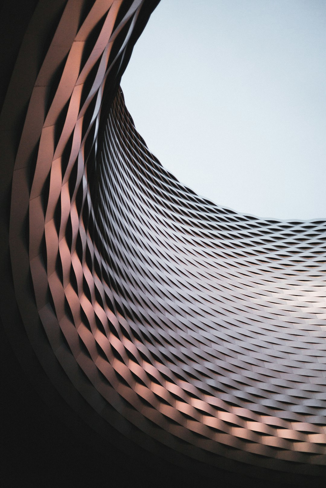 A photograph of an abstract building with copper metal strips, a circular design, a curved surface, looking up from the ground at dusk. The photograph was shot on a Sony Alpha A7 III in the style of an abstract expressionist painting.