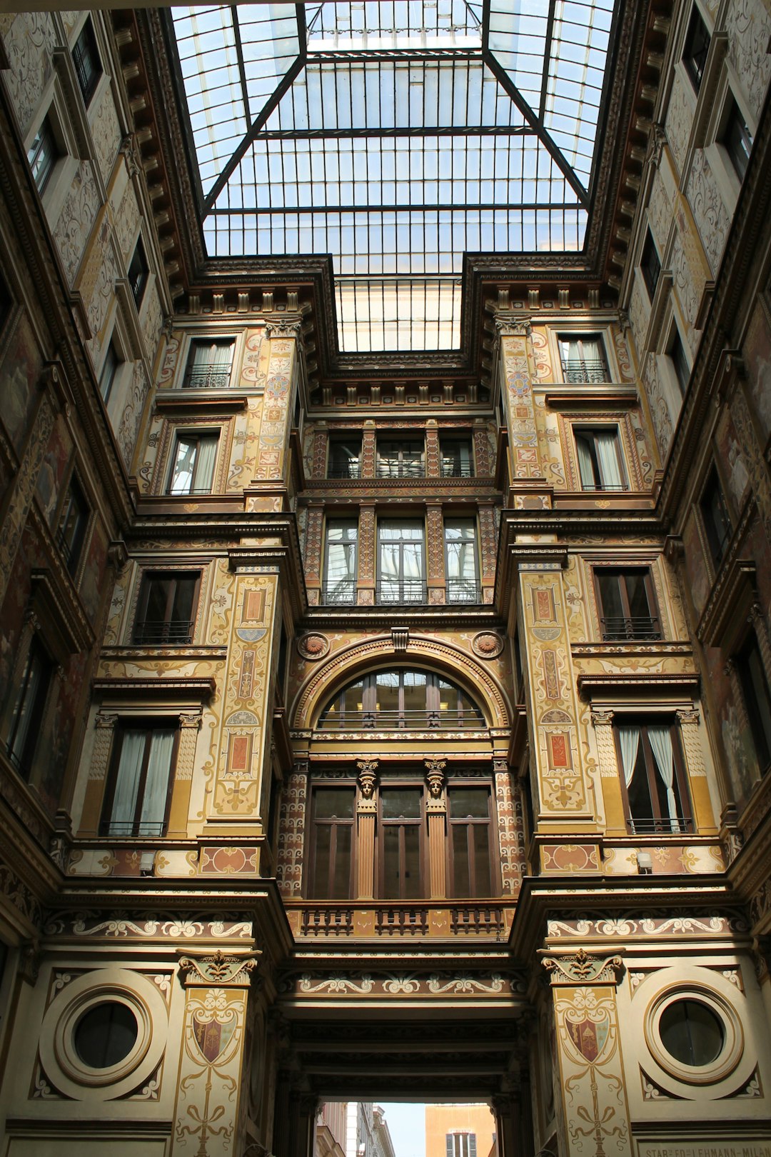 The photograph shows the inside of an old building in Italy with ornate details and a glass roof above, showing its tall ceilings and large windows. The architecture is elegant and intricate, with decorative elements such as stucco work on walls and columns, and painted designs that add to its visual appeal. In front there is another open space where you can see other buildings or street scenes through the window. It seems like part of a busy city center. The photograph appears to be in the style of a Renaissance artist, capturing ornate architectural details with natural light. –ar 85:128