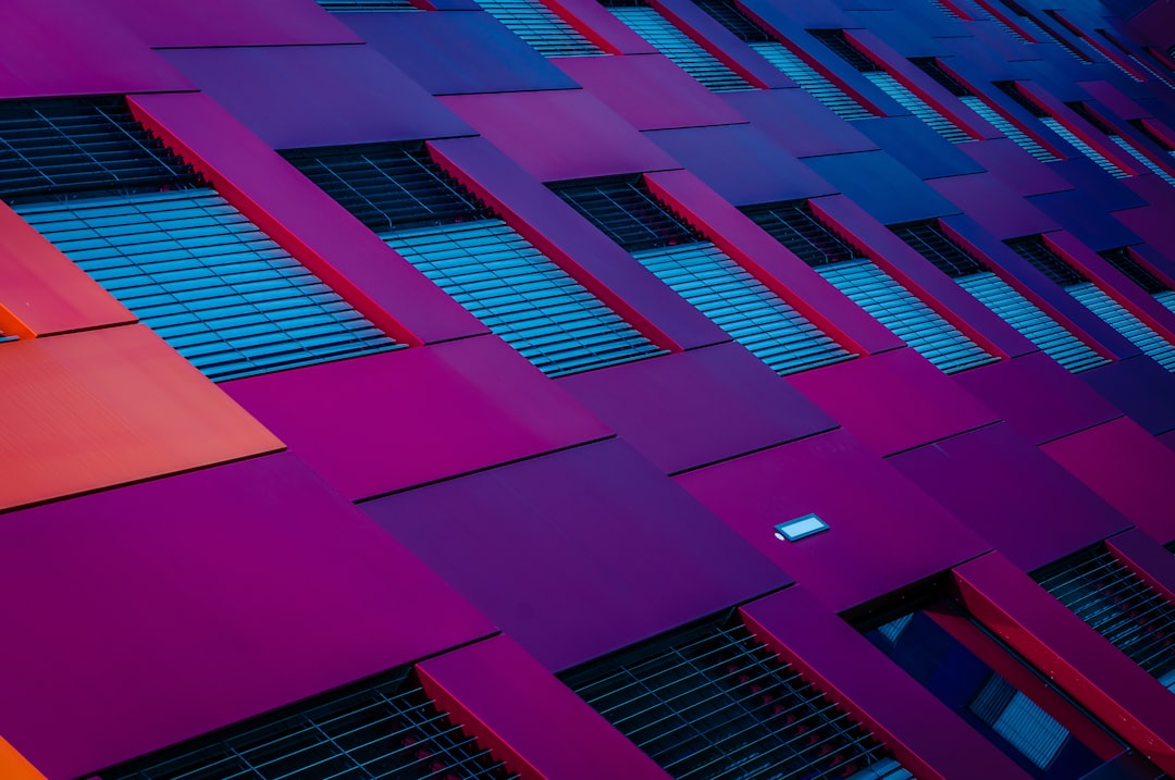 A photograph of an abstract building facade with vibrant red and purple colors, geometric shapes, and windows with blue screens, captured from the outside using a wide-angle lens at sunset. The image was taken with a Sony Alpha A7 III camera and shows a close-up view of architectural details in the style of geometric abstraction.