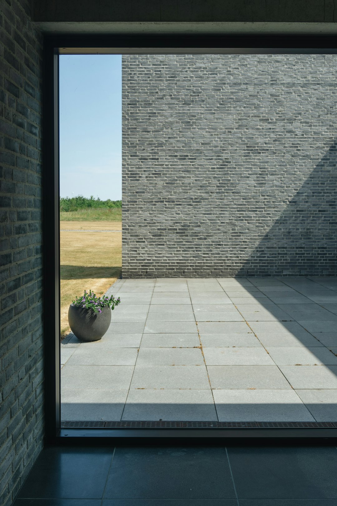 An open wall with grey brick and a concrete floor overlooks a field in Denmark. There is a large window on one side of an outdoor room that has a stone slab surface as flooring. A pot plant sits next to it. The sky outside shows blue skies. Photography. Minimalistic architecture photographed in the style of Architectural photography. –ar 85:128