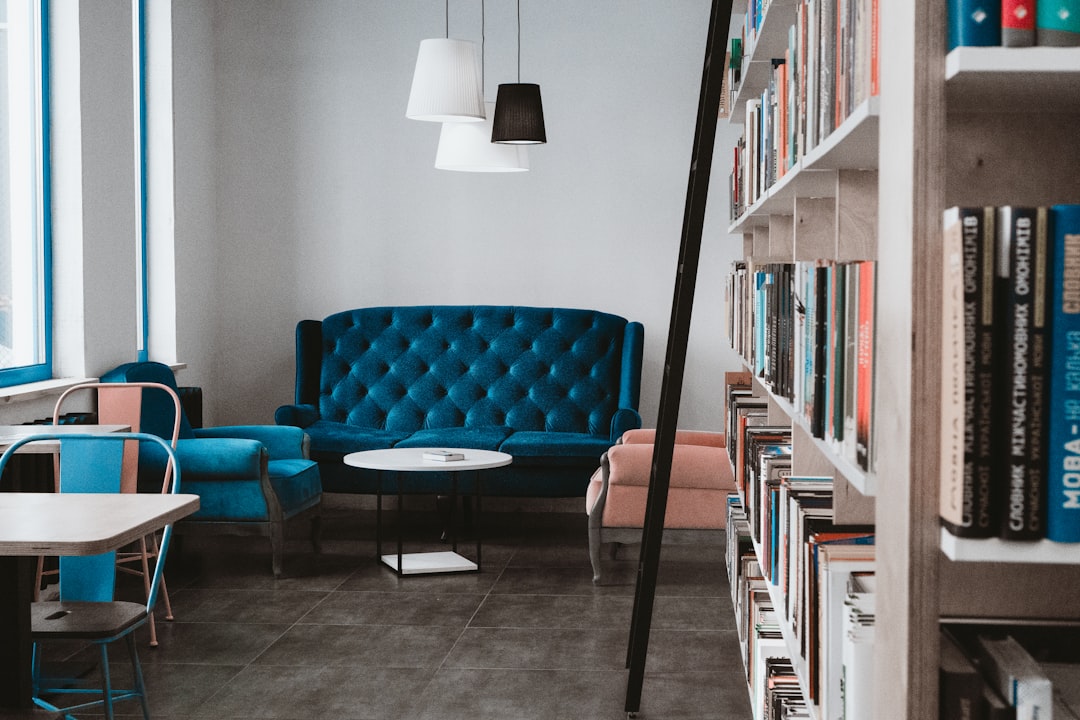 A modern library with blue sofas, white walls and bookshelves on the right side of the picture. In the center there is an empty table in front of the sofa. On one wall there is a hanging lamp above a coffee table. There is also another sofa behind a black ladder. The photo was taken from a wide angle. The photo was taken with a Canon EOS camera using a macro lens. –ar 128:85