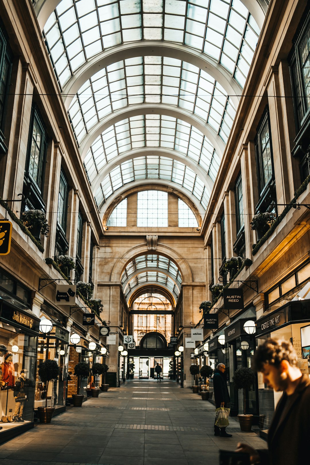 A photo of the interior of an old Victorian shopping center with a glass roof, surrounded by shops and fashion stores. The scene is bustling with people walking around. In the background there’s another street with buildings and a traffic light. Use a Sony α7 III camera with an 85mm lens at F/4 aperture setting to capture the subject in sharp focus against their urban surroundings. Use soft lighting to create gentle shadows on your face in the style of Sony. –ar 85:128