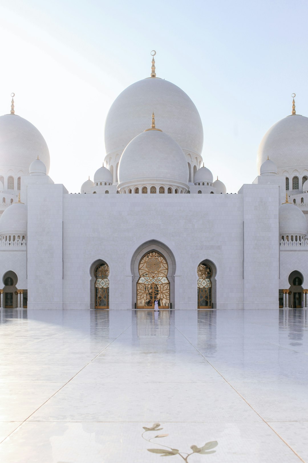 A wide angle photo of the white and golden dome on Shepard Kahane’s mosque in Abu Dhabi, shot from outside with a clear sky. The polished marble floor is shown, with an open door leading to another building with gold details. A man wearing a thobe is walking inside it in the style of hyper realistic, professional photography lit by natural light. –ar 85:128