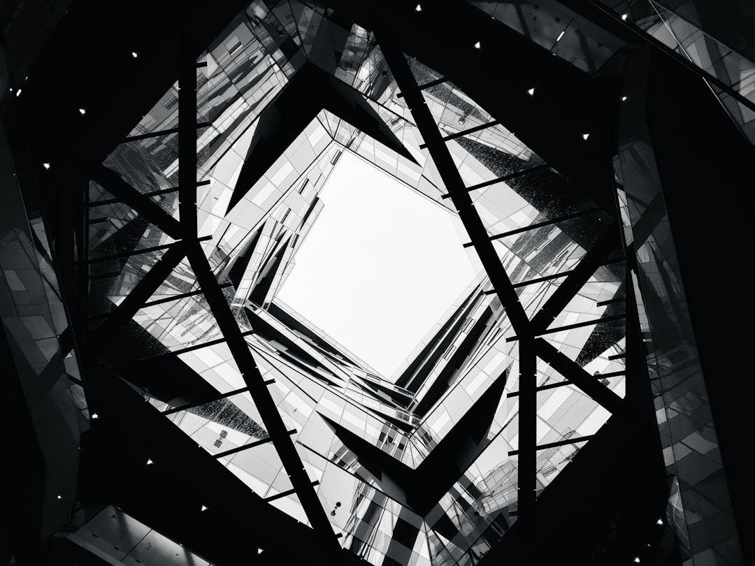 A stunning black and white photo shows an abstract geometric architecture. The glass structure has angular geometry that forms the interior space of a building. Looking up from below through a skylight, light can be seen shining down on all sides, creating a symmetrical pattern of highlights in contrast to dark shapes. The scene is captured from inside the tall, tower-like skyscraper in the style of an abstract geometric style.