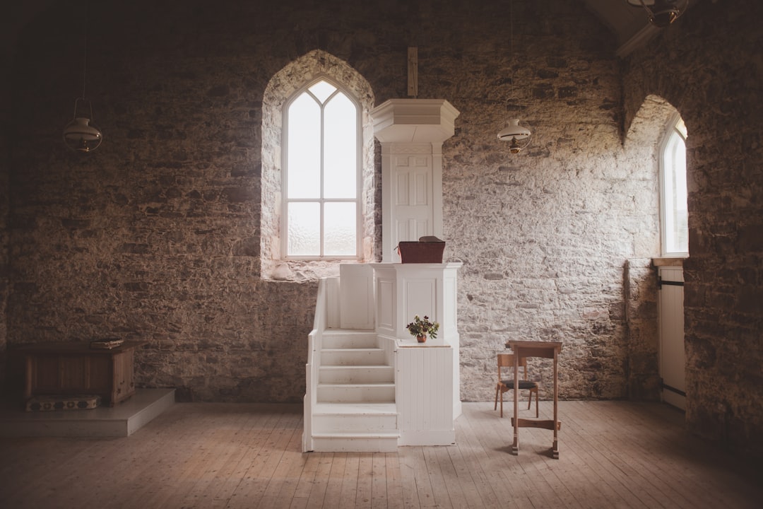 A photo of an empty church hall with white wooden stairs leading up to the pulpit, a small side table at one end holding fresh flowers and candles, tall arched windows casting soft light from outside, a stone wall behind it, creating a cozy atmosphere for a wedding photographer’s photoshoot. –ar 128:85