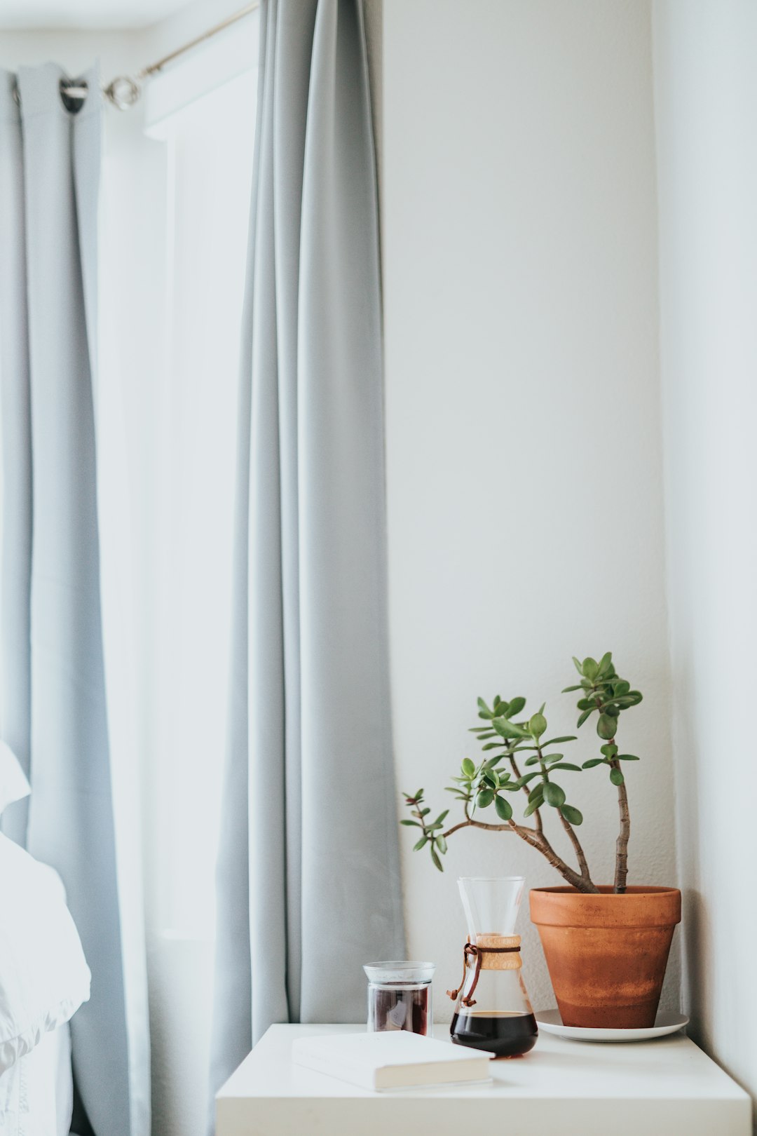 A close up photo of the side table in the bedroom with grey curtains, a coffee pot and a small plant on top, in a minimalist style with natural light and a white wall, shot in the style of Canon EOS R5 at F2, ISO400 and stylized at 750. –ar 85:128