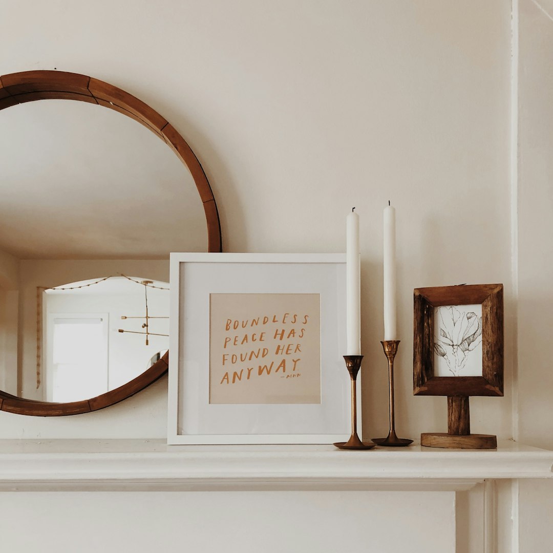 A simple, yet elegant display of the quote on an A4 frame with warm beige and white tones, placed above a minimalist fireplace mantel adorned by two wooden candlesticks, a vintage mirror reflecting the light from a nearby window, creating soft shadows that accentuate its text against a plain cream wall background. The overall atmosphere is one of tranquility and simplicity, capturing both depth in design and warmth in ambiance. golden hour