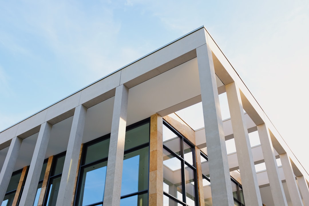 A modern building with columns and windows, featuring an exterior made of white concrete and beige stone, under the clear blue sky on a sunny day. The architecture is contemporary and sleek, showcasing elegant lines and minimalist design elements in the style of modern architects. –ar 128:85