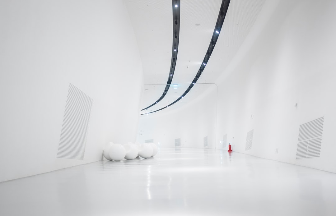 A white room with long curved LED strips on the ceiling, a few large stone spheres and minimalistic wall panels on one side of an empty space, a person in red  standing at the far end of the hall. Minimalist architecture. Canon EOS camera, 35mm lens, f/2. –ar 64:41