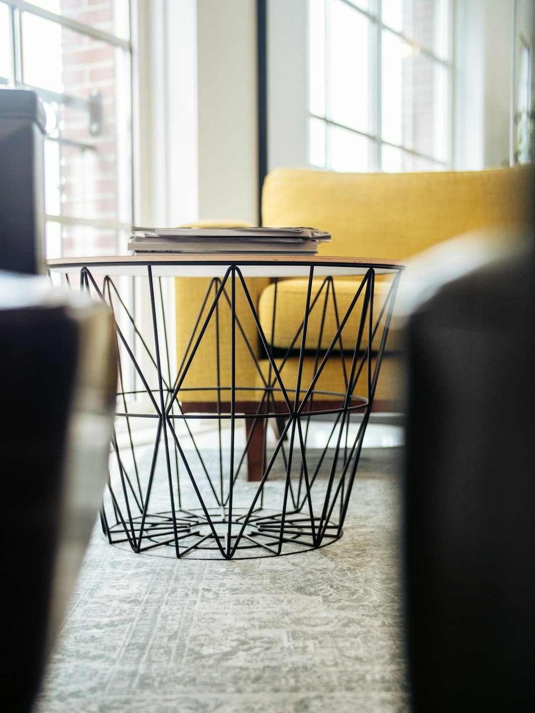 A closeup shot of an industrial wire frame coffee table, placed in the corner with yellow sofas and black chairs, books on top of it. The background is a bright office space with large windows that provide natural light. This photo was taken using a Canon EOS R5 camera, focusing on detail to highlight intricate patterns and textures of the metal wires used for creating geometric shapes. Soft lighting creates a warm atmosphere, adding depth to the scene. This photo was taken in the style of capturing intricate details and textures.