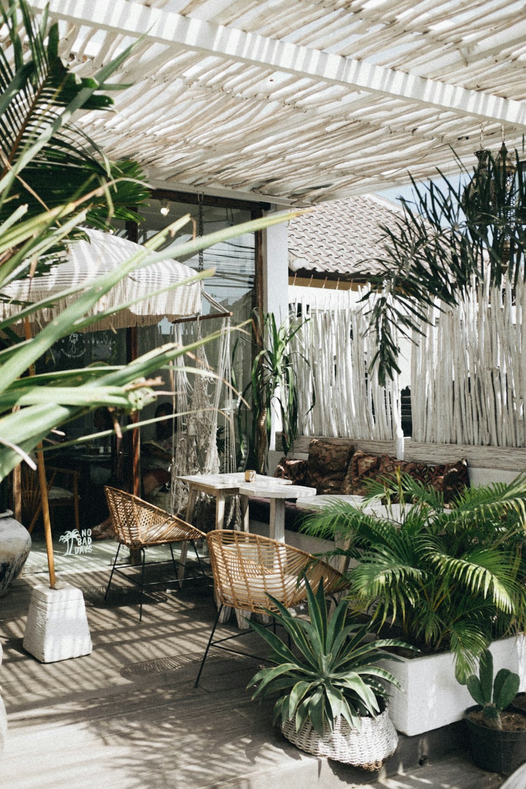 A photo of an open air cafe with white wood slats on the roof, surrounded by plants and wicker chairs, taken from above, shot in natural light, in the style of unsplash photography.