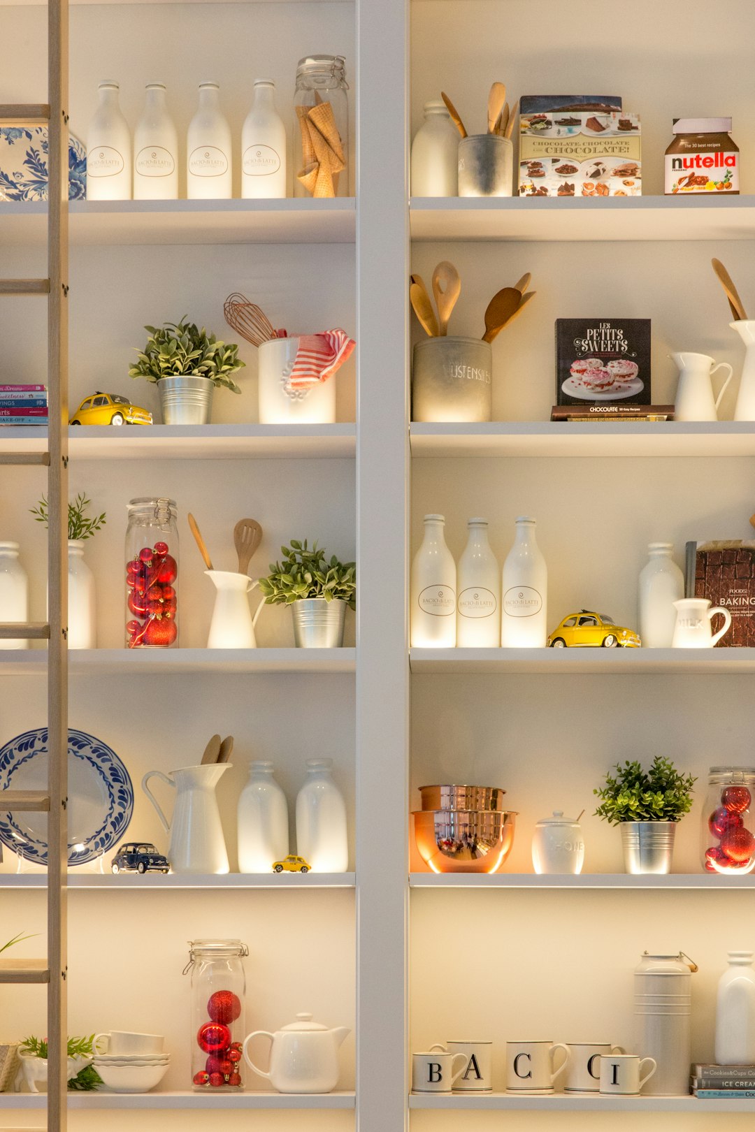 White shelves with backlight, a closeup of the top shelf in an open kitchen. The left side is decorated with various home appliances and decorations such as coffee mugs, pots, wooden cooking utensils, plates, candles, red fruits on ceramic vases, white bottles of “Nincture” milk brand, colorful books, small yellow car toy figurines, white glass jars for cream or oil, and a black book cover that says ‘Blowing’ in blue letters in the style of B. –ar 85:128