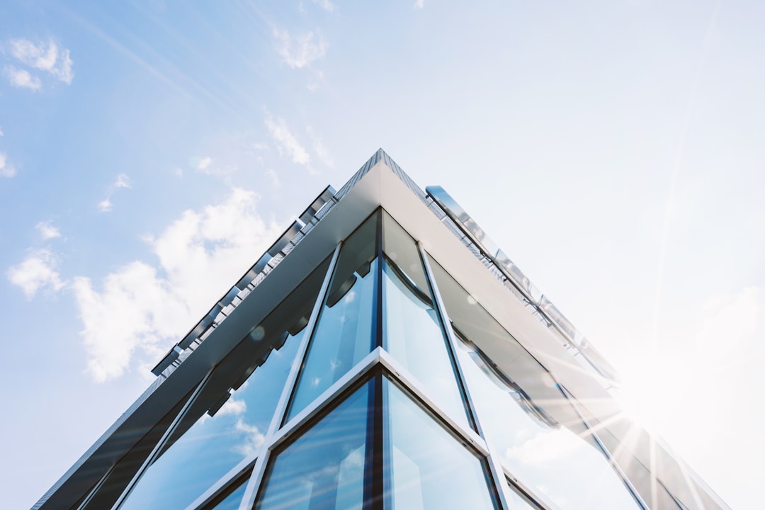 Modern office building exterior with blue sky and clouds, low angle view of the corner of glass facade, sunny day. wide shot, Use a Canon EOS R5 camera at F20 aperture setting to create soft focus on background elements. The sunlight is streaming through, creating light rays that add depth and dimension to the scene. High resolution, photo realistic quality –ar 128:85