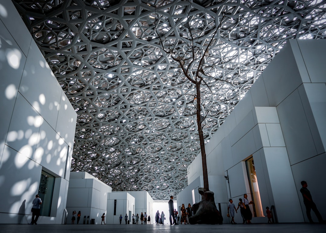 Inside the branch of the Louvre in Abu Dhabi, a steel and glass ceiling with a tree inside. Many people walking around. A wide shot of the architecture, photography in the style of architecture. –ar 128:91