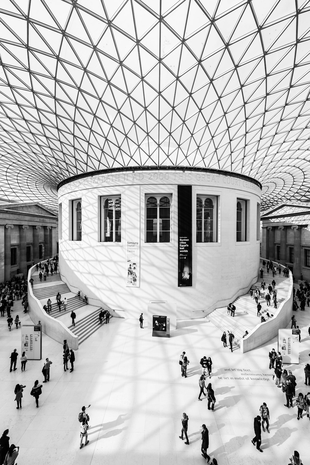 The British Museum in London has a white interior with black geometric patterns on the ceiling. Busy and bustling crowds of visitors surround it. Black and white photography captures the natural lighting and interior in the style of architectural photography using a Hasselblad X2D with a wideangle lens and 35mm f/4L lens. –ar 85:128